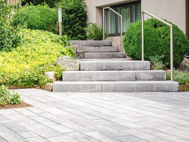 Gepflasterte Terrasse mit Steintreppe, Geländer und begrüntem Beet vor einem Hauseingang.