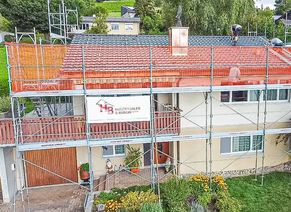 Ein Einfamilienhaus mit rotem Ziegeldach ist rundum mit Gerüst und orangem Schutznetz eingerüstet.
