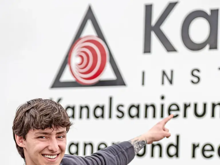 Ein junger Mann in Arbeitskleidung lacht und zeigt mit beiden Händen auf einen Firmenschriftzug.