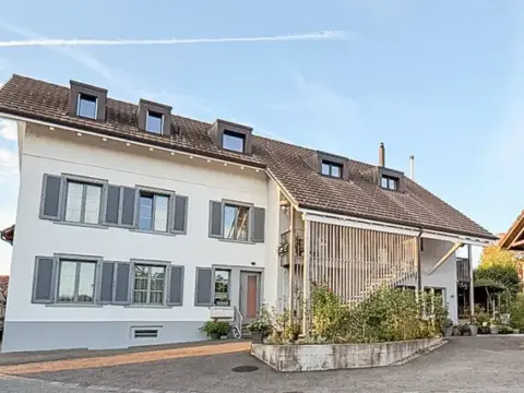 Umgebautes Bauernhaus mit weisser Fassade, Sprossenfenstern und angrenzender Holzscheune unter blauem Himmel.