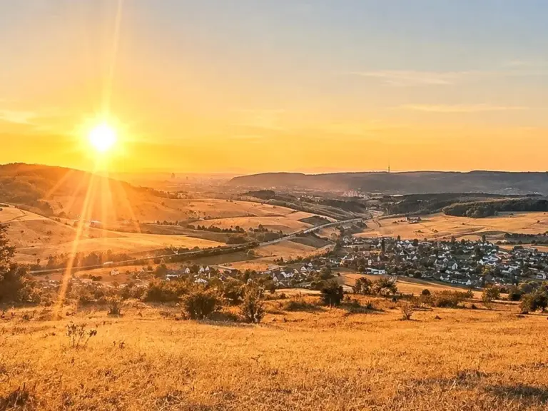 Sonnenuntergang über einem Tal mit Dorf, Feldern und Hügeln, im Vordergrund goldene Wiese.