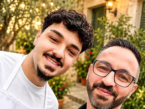 Zwei lächelnde Männer mit Schürzen posieren für ein Selfie vor einem Innenhof mit Lichterketten.