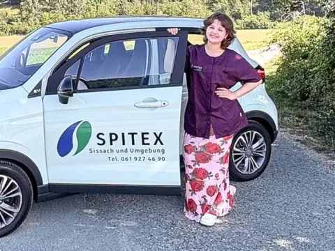 Eine Frau lehnt lächelnd an einem weissen Spitex-Auto vor einer Hügellandschaft.