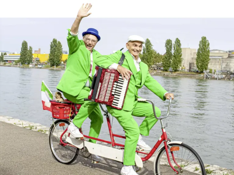 Zwei ältere Männer in grünen Anzügen fahren auf einem roten Tandem am Flussufer, einer winkt, der andere spielt Akkordeon.