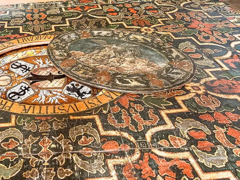 Bemalte Tischplatte mit ornamentalem Rankenmuster, lateinischer Inschrift und rundem Landschaftsmedaillon mit Wappen.