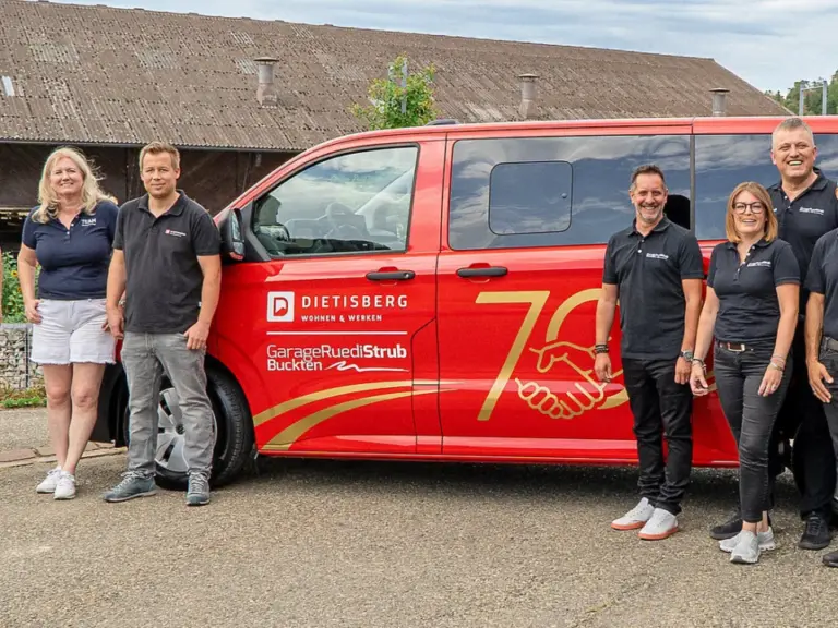 Acht Personen in Poloshirts stehen vor einem roten Kleinbus mit Werbeaufdruck vor einem Bauernhof.