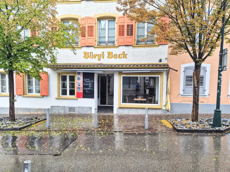 Fassade einer Bäckerei mit Schriftzug „Bürgi Beck“ an einer regennassen Strasse mit herbstlichen Bäumen.