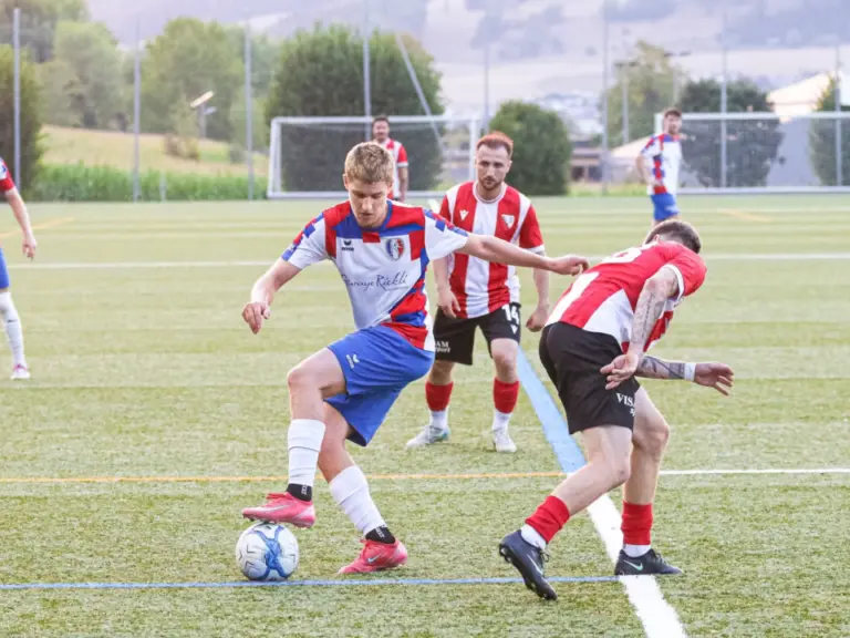 Fussballspieler kämpft am Ball gegen einen Gegenspieler, weitere Spieler im Hintergrund auf dem Rasen.