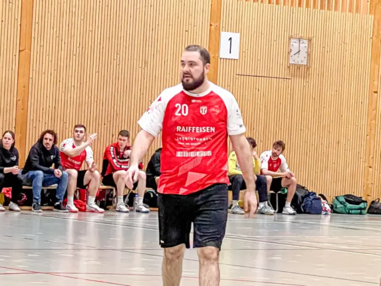 Ein Handballspieler im roten Trikot mit Nummer 20 geht über das Spielfeld, im Hintergrund sitzen Zuschauer.