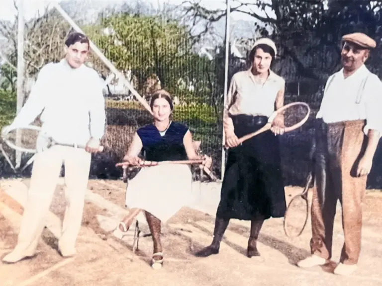 Vier Personen in altmodischer Kleidung stehen mit Tennisschlägern auf einem Sandplatz.
