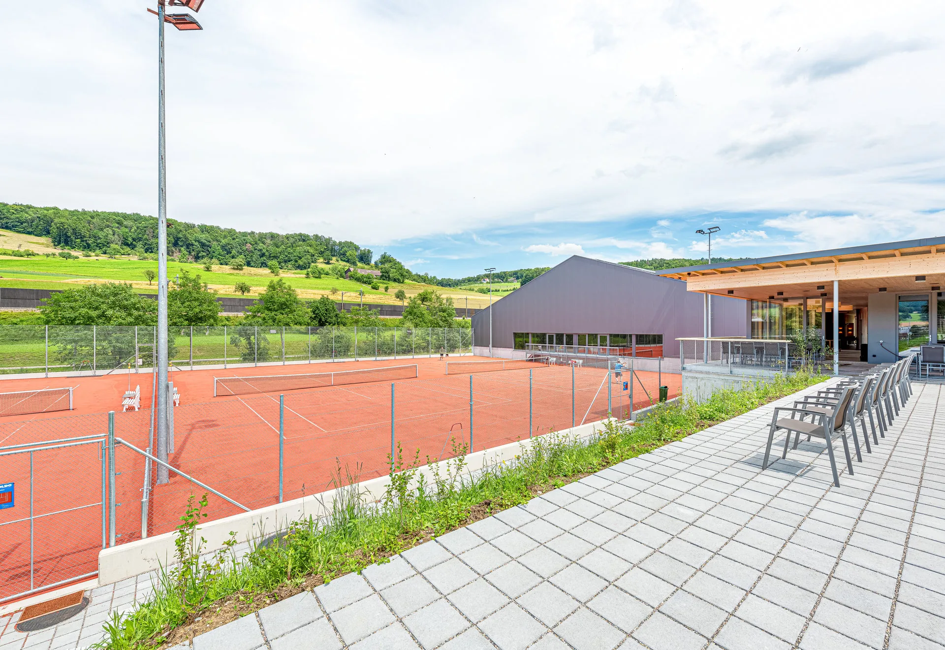 Tennisplatz mit Flutlicht neben einem modernen Vereinsgebäude und Terrasse mit Stühlen, umgeben von grünen Hügeln.