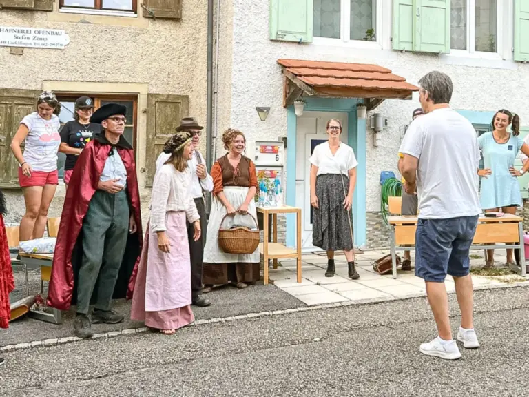 Menschen in historischen Kostümen stehen vor Häusern in einer Dorfstrasse, ein Mann filmt sie.