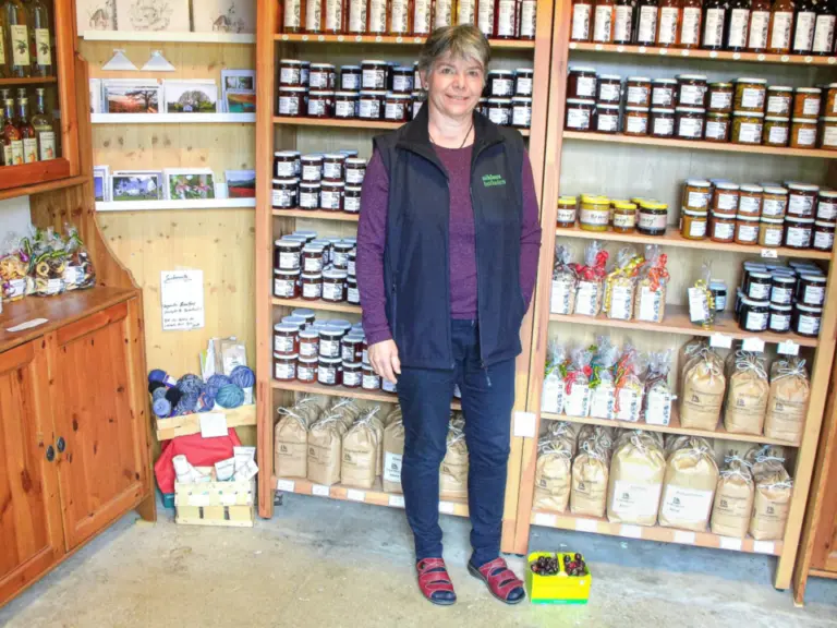 Eine Frau steht in einer Weste in einem Hofladen vor Regalen mit Marmeladengläsern und Gebäcksäcken.