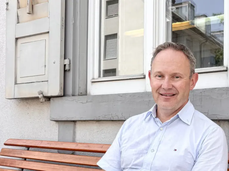 Ein Mann im hellblauen Hemd sitzt lächelnd auf einer Holzbank vor einem Fenster mit Fensterläden.