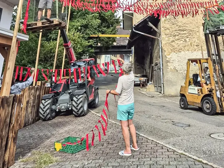 Eine Person spannt rote Girlanden über eine Dorfstrasse, im Hintergrund helfen Männer auf einer Hebebühne.