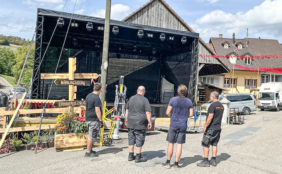 Drei Männer in Arbeitskleidung stehen vor einer schwarzen Bühne im Freien und betrachten diese.