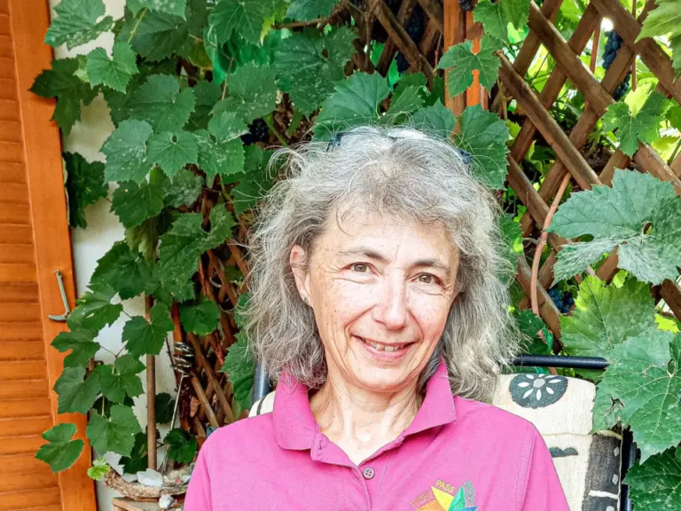 Eine lächelnde Frau sitzt im Garten auf einem Stuhl und hält ein türkises Heft mit der Aufschrift Ferienpass.