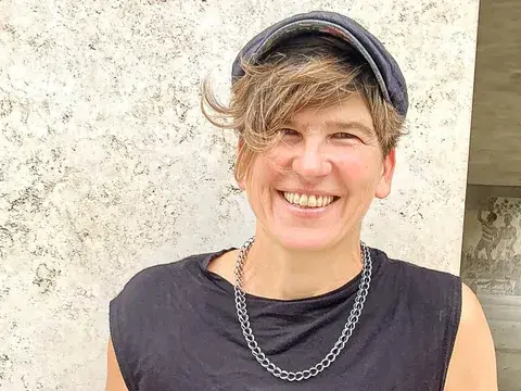 Eine lachende Frau mit kurzen Haaren und Cap trägt ein dunkles Top und eine Kette vor einer Steinwand.
