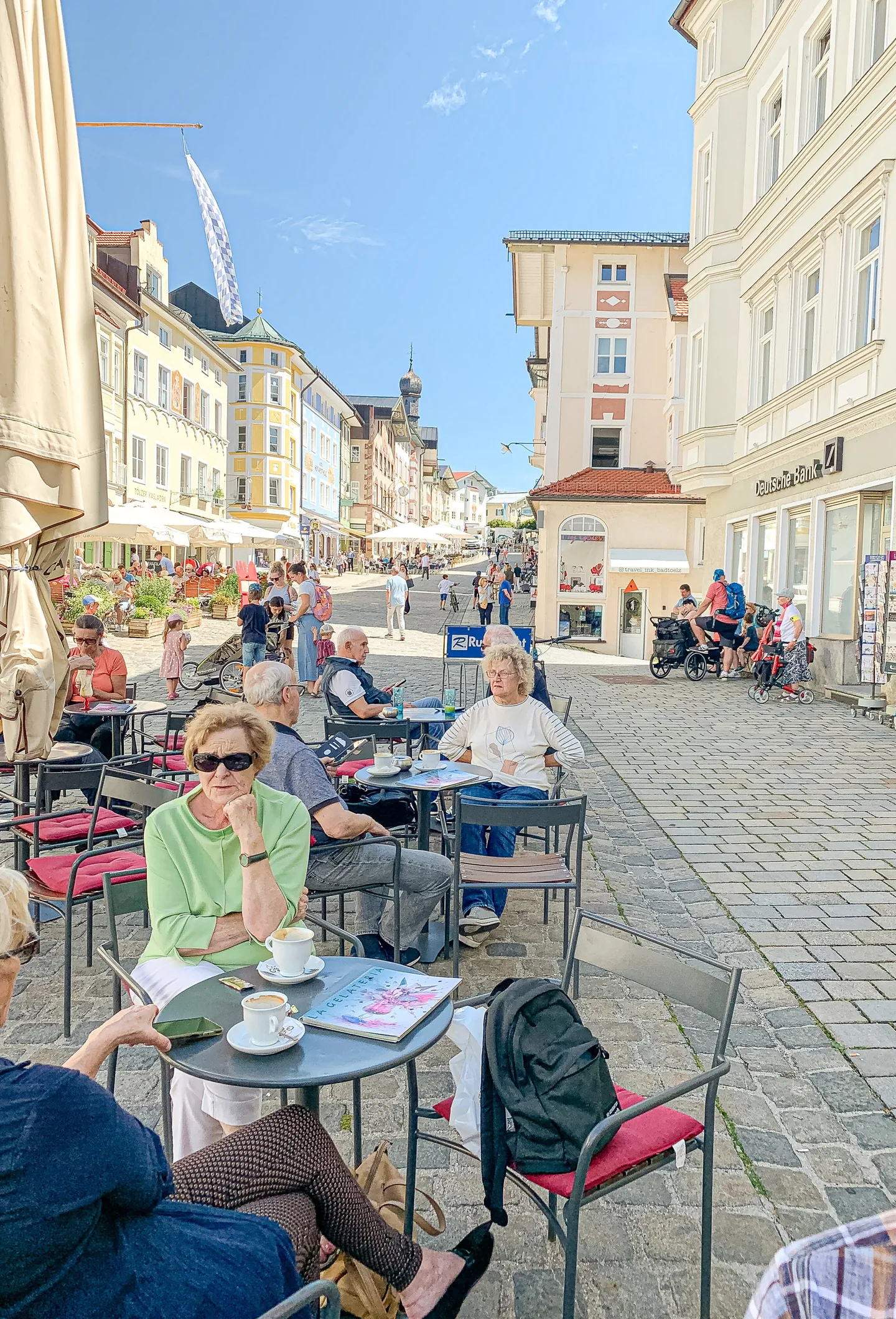 Menschen sitzen an Cafétischen in einer sonnigen Fussgängerzone mit historischen Häusern.