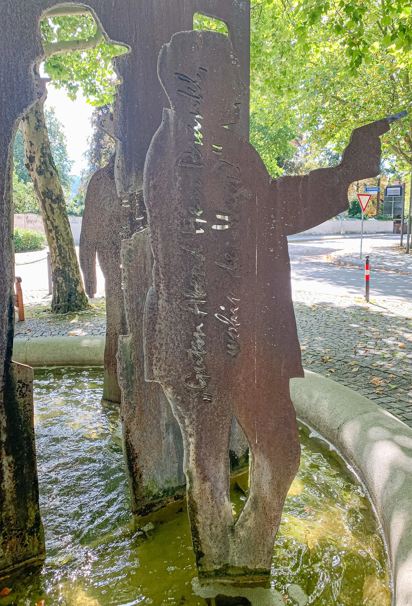 Rostige Metallsilhouetten von Männern mit Hüten in einem grünlichen Wasserbecken, umgeben von Bäumen.