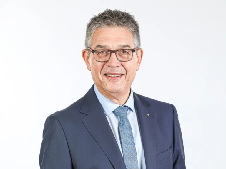 Ein älterer Mann mit Brille und grauem Haar im dunkelblauen Anzug lächelt vor hellem Hintergrund.