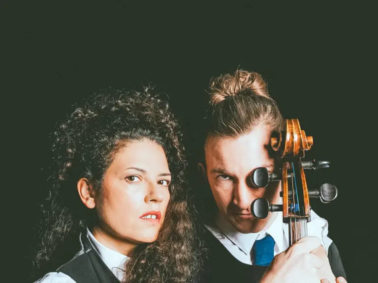 Eine Frau mit lockigem Haar hält ein Keyboard, ein Mann mit Dutt hält ein Cello, beide blicken in die Kamera.