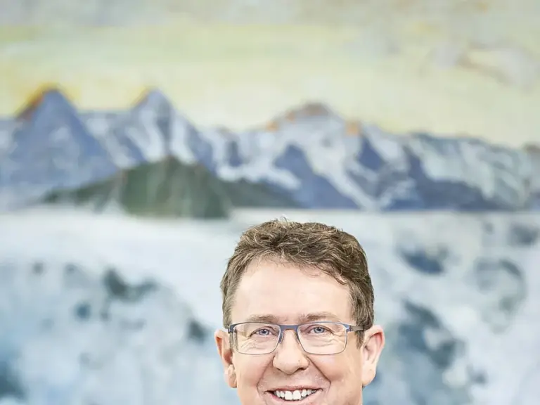 Ein lächelnder Mann mit Brille und blauem Anzug samt Krawatte steht vor einem Gemälde mit Berglandschaft.