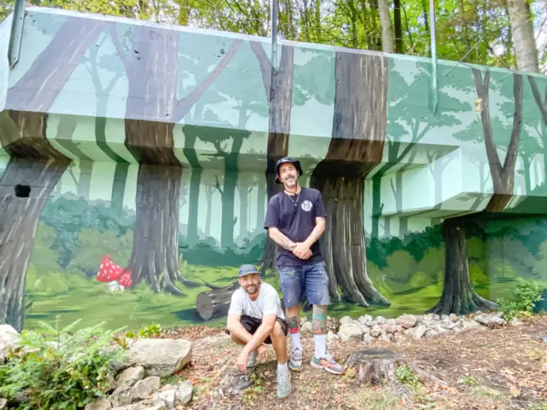 Zwei Männer stehen und knien vor einer bemalten Wand mit einem Waldmotiv im Freien.
