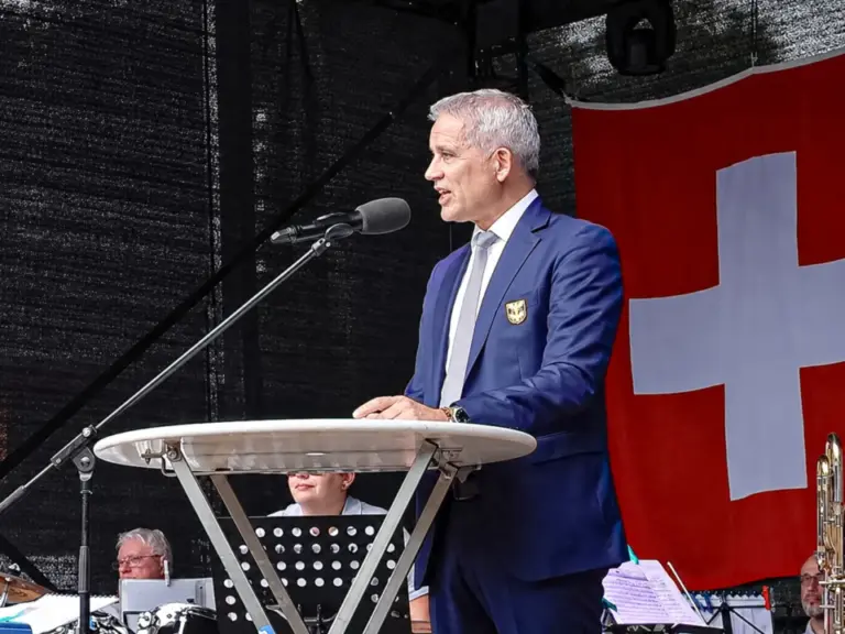 Ein Mann im dunkelblauen Anzug spricht an einem Mikrofon auf einer Bühne vor einer Schweizer Fahne.