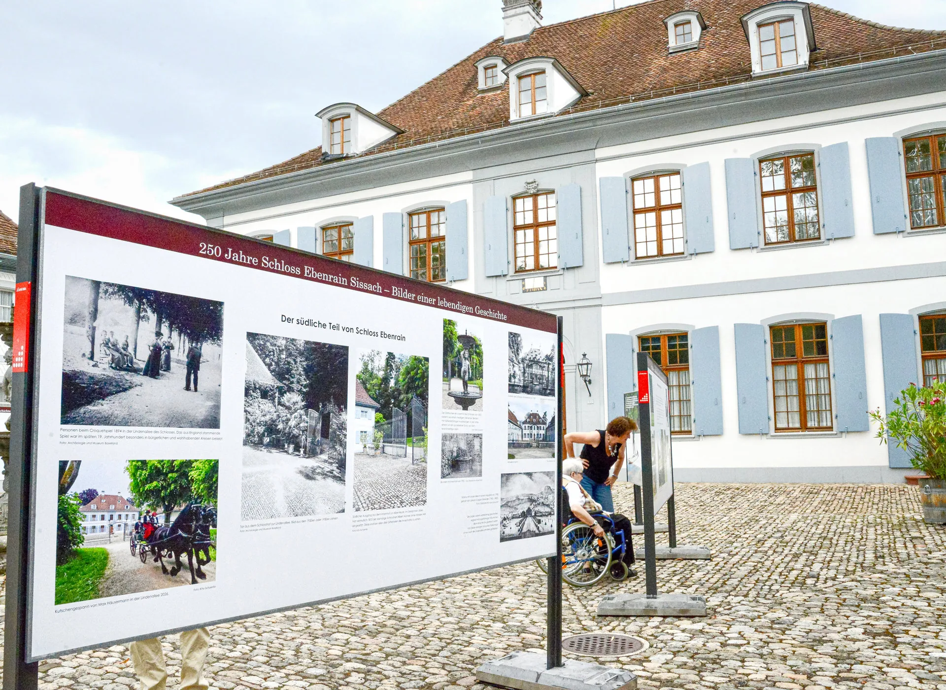 Eine Ausstellungstafel mit historischen Fotos steht vor einem Schloss, im Hintergrund hilft eine Person einer im Rollstuhl sitzenden Person.