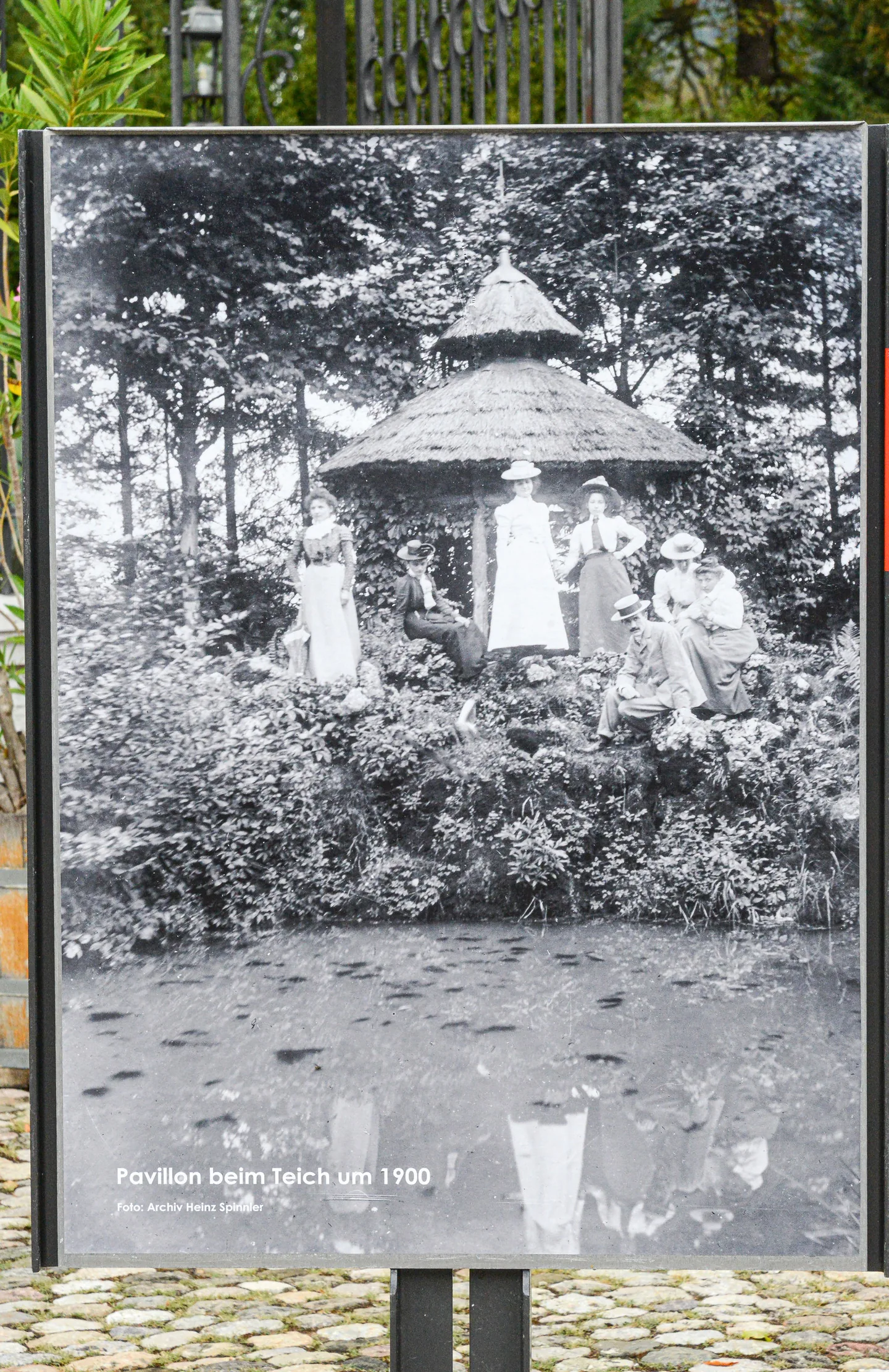Informationstafel mit historischem Schwarzweißfoto eines Pavillons mit Menschen in altmodischer Kleidung am Teich.