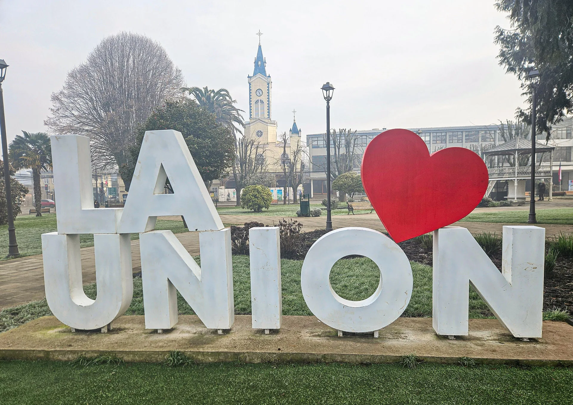 Grosse weisse Buchstaben „LA UNION“ mit rotem Herz auf einer Grünfläche, im Hintergrund eine Kirche.