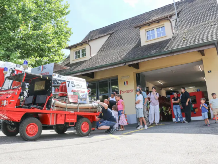 Menschen stehen vor einem Feuerwehrhaus, im Vordergrund ein alter roter Feuerwehr-Anhänger mit Schlauch.