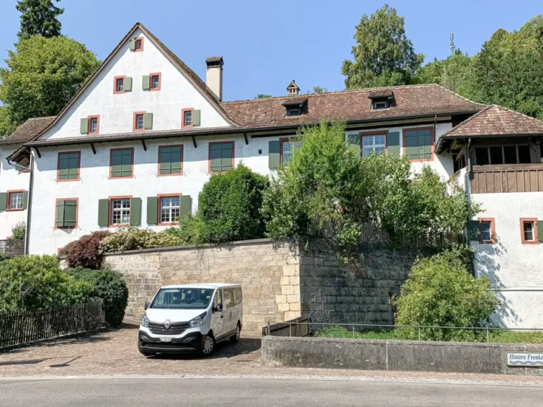 Grosses, altes Pfarrhaus mit weisser Fassade und Klappläden, davor eine Steinmauer und ein parkiertes Auto.