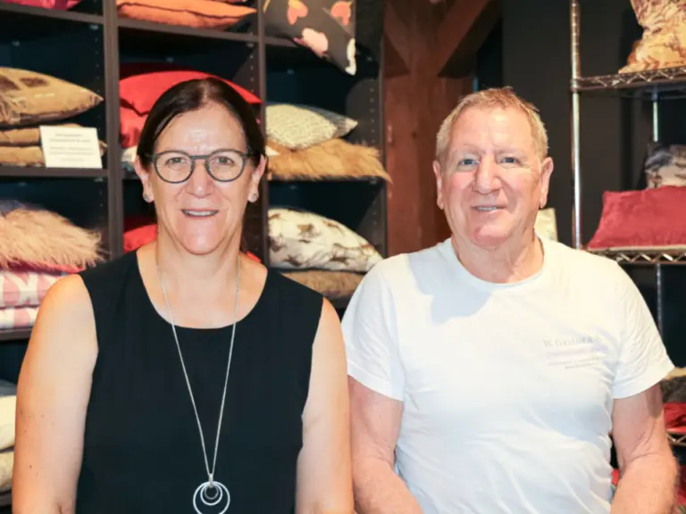 Eine Frau mit Brille und Kette und ein älterer Mann stehen lächelnd in einem Laden voller Kissen.