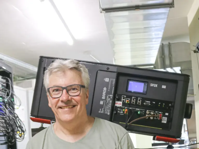 Ein lächelnder Mann mit Brille sitzt auf einem Hocker in einem Technikraum voller Geräte und Kabel.