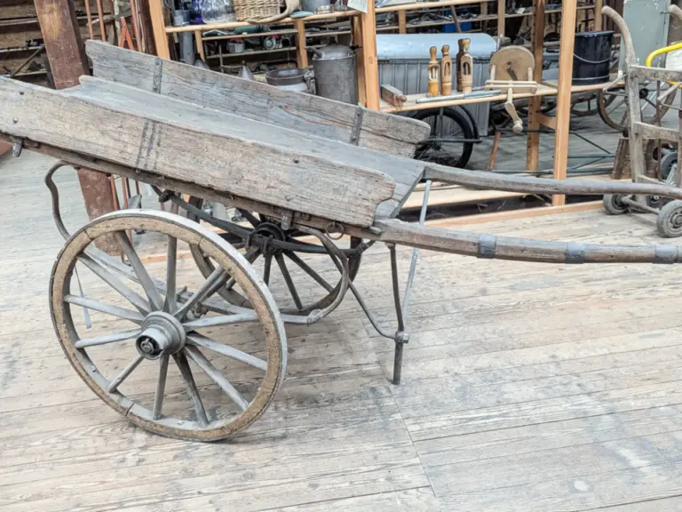 Ein alter zweirädriger Holzhandwagen mit einer Deichsel steht auf einem Holzboden in einer Werkstatt.
