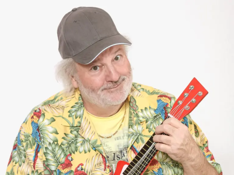 Ein älterer Mann mit grauer Kappe und Hawaiihemd sitzt lächelnd und spielt eine rote Ukulele.