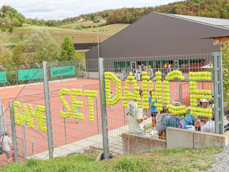 Tennisanlage mit Zaun, an dem aus Tennisbällen die Worte „Game Set Danke“ gestaltet sind, im Hintergrund Hügellandschaft.