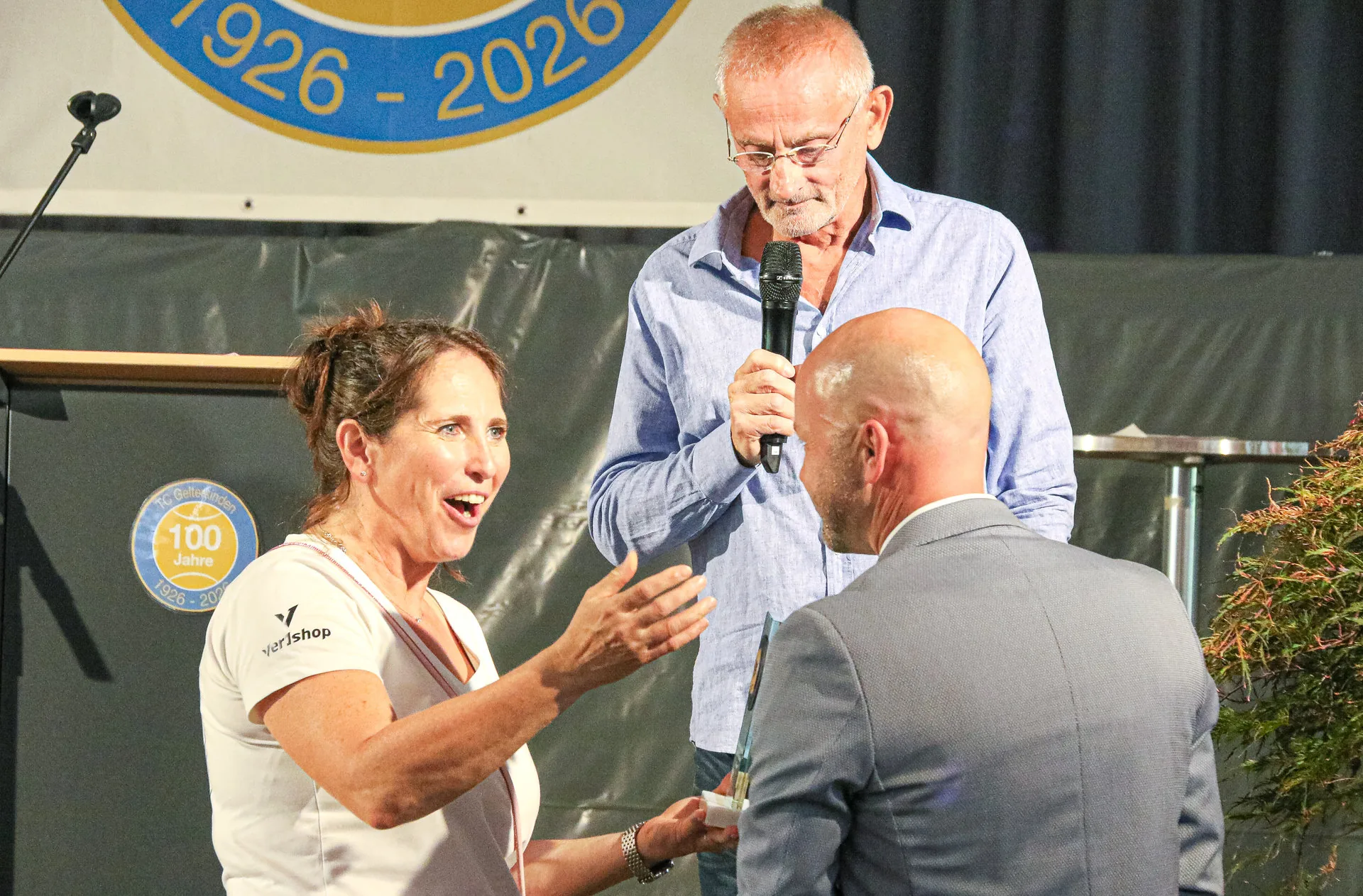 Eine Frau gestikuliert im Gespräch mit einem Mann im Anzug, während ein Mann mit Mikrofon dahinter steht.