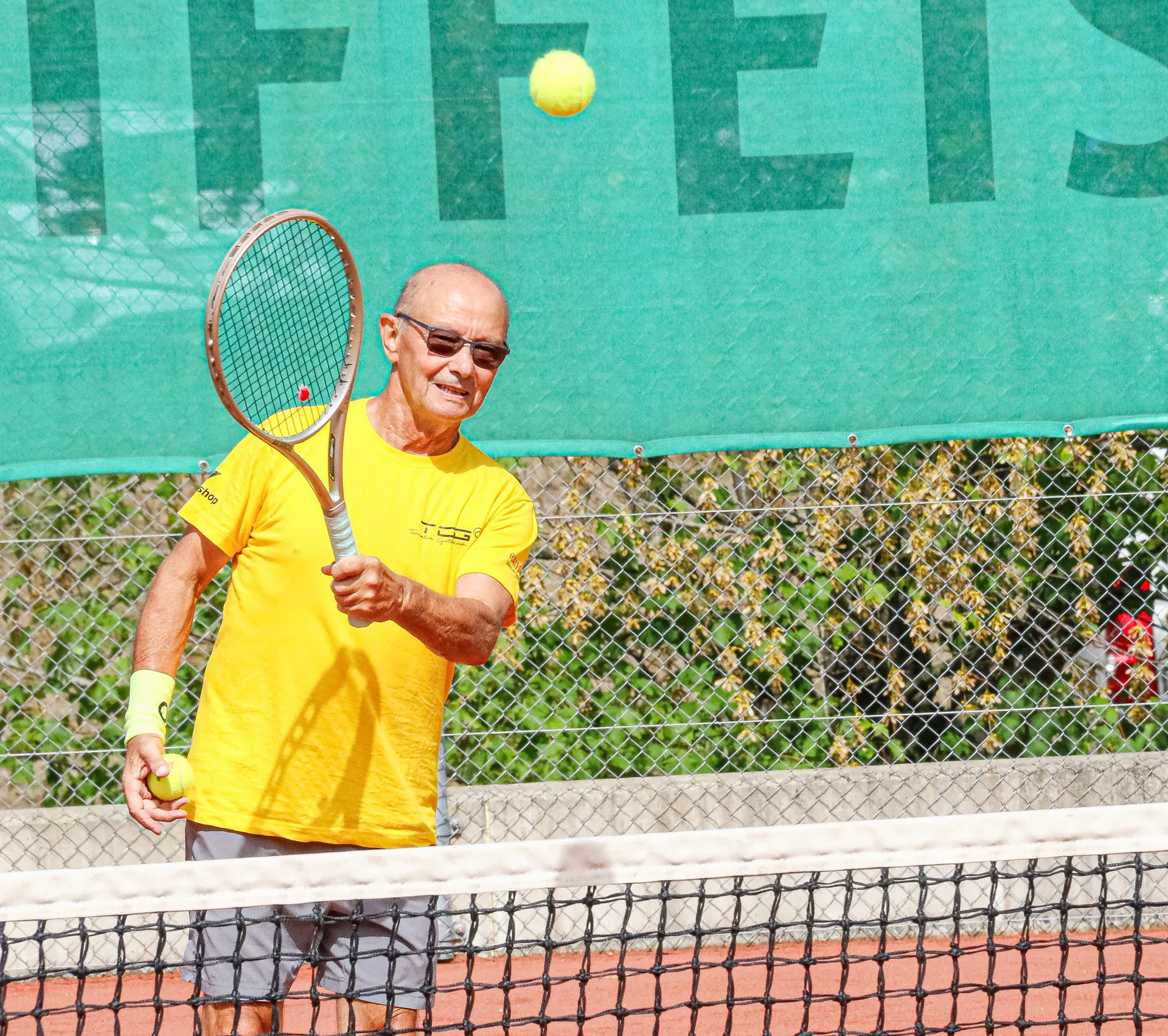 Älterer Mann in gelbem Shirt spielt mit Schläger Tennis auf einem Sandplatz vor grünem Werbebanner.