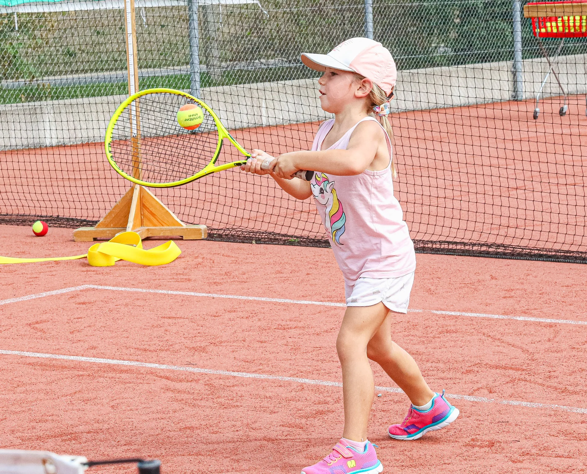 Ein kleines Mädchen mit Cap schlägt auf einem Tennisplatz mit dem Schläger nach einem Ball.