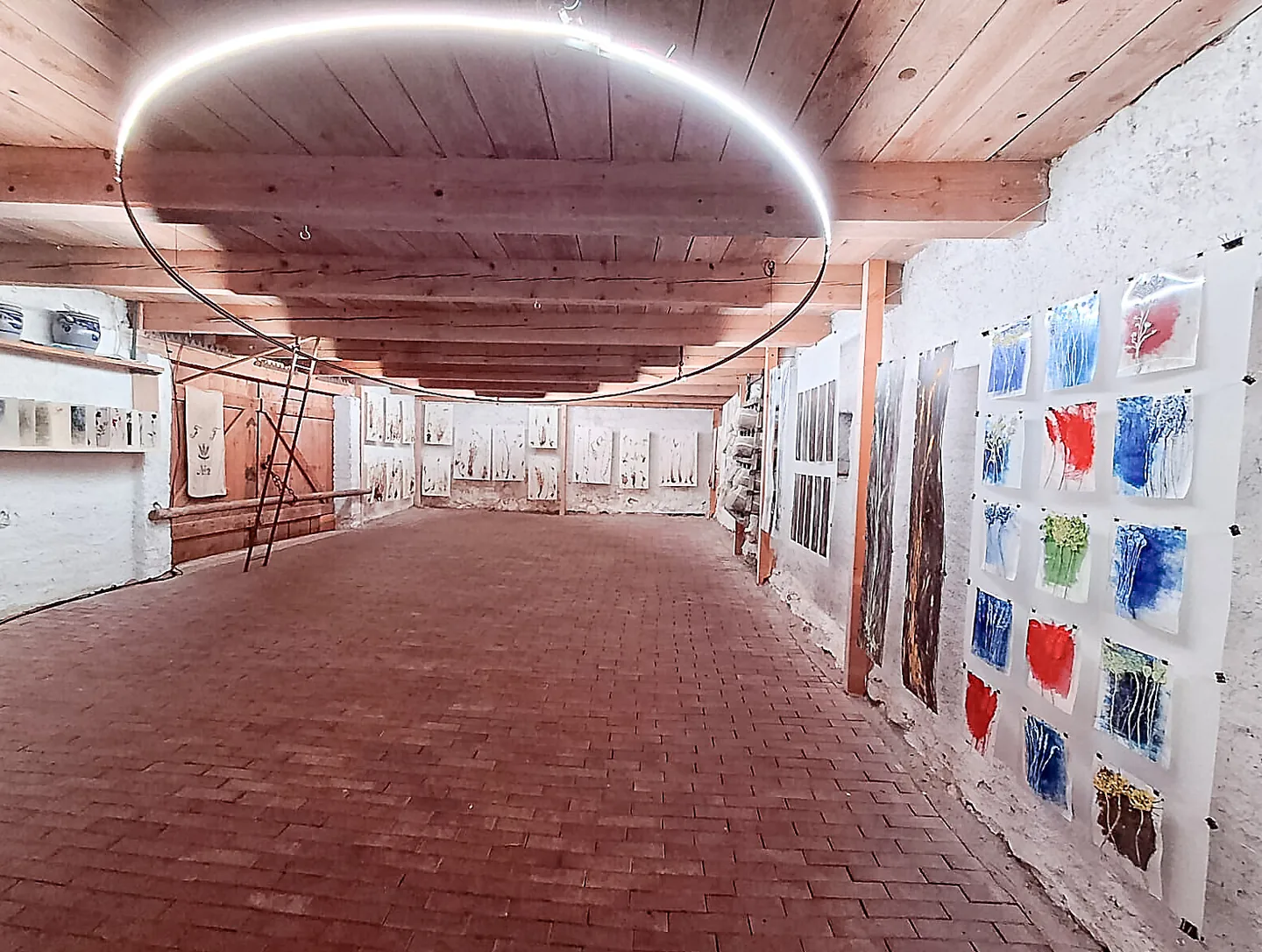 Ausstellungsraum mit Ziegelboden, Holzdecke und Kunstwerken an weissen Wänden, darüber ein leuchtender Ring.