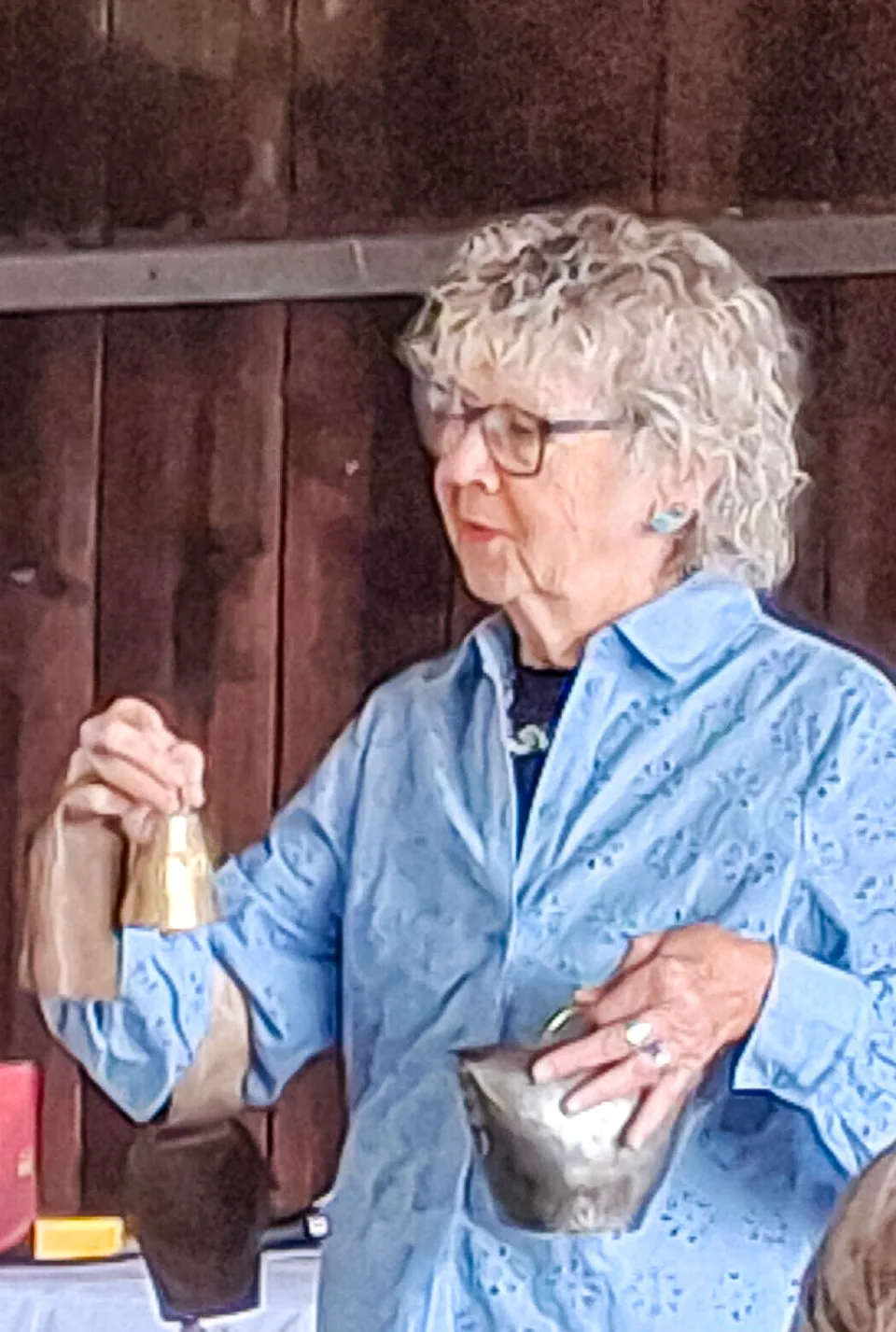 Ältere Frau mit lockigem weissem Haar und Brille hält eine Glocke und eine Schale in den Händen.