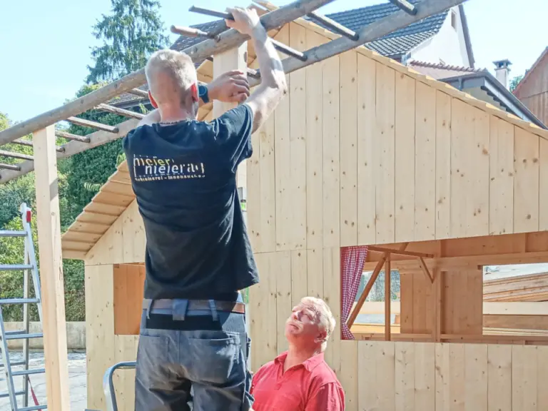 Zwei Männer arbeiten an einem Holzhaus, einer steht auf einer Leiter und montiert Dachlatten, der andere schaut hoch.