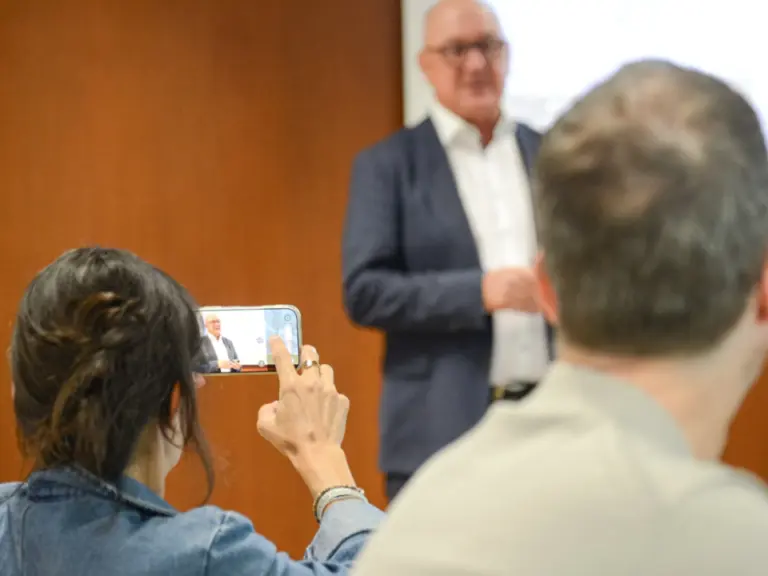 Ein älterer Mann im Anzug spricht vor Publikum, eine Zuhörerin filmt ihn mit dem Smartphone.