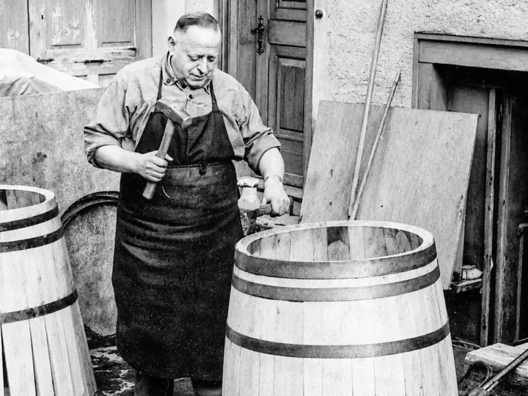 Ein Mann in Schürze bearbeitet mit Hammer ein hölzernes Fass im Hof.