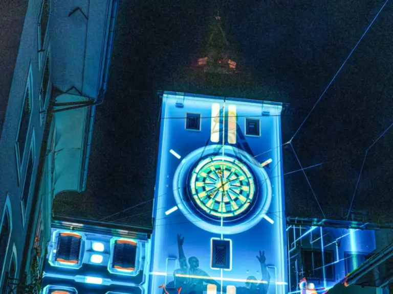 Ein beleuchteter Stadtturm mit Uhr wird nachts bunt illuminiert, davor stehen zahlreiche Menschen.