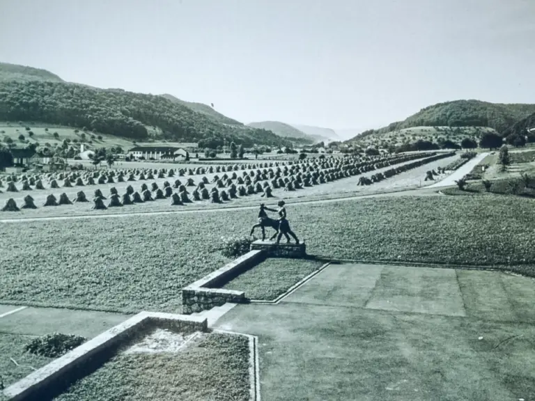 Schwarzweiss-Aufnahme einer Statue auf Sockel, dahinter Reihen von Feldern, Hügel und ein Gebäude.