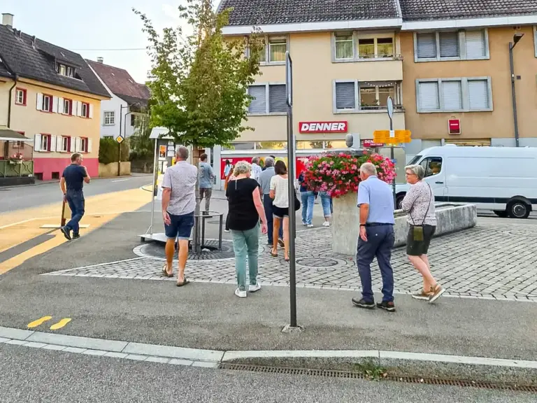 Mehrere Personen stehen und gehen über einen Dorfplatz mit Blumenschmuck vor einem Denner-Laden.